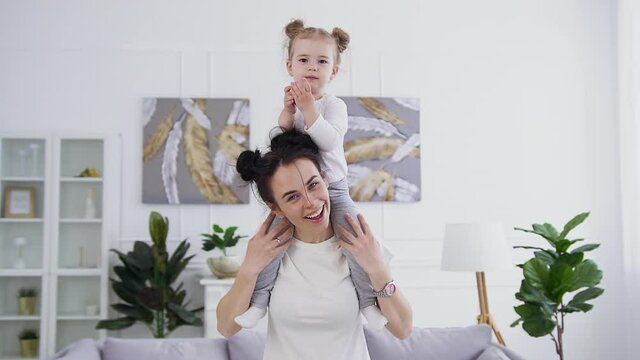 Attractive happy smiling young woman with funny hairstyle holding on shoulders her satisfied little daughter ,posing on camera at home