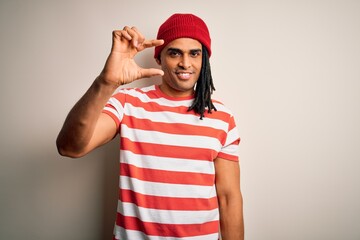 Young handsome african american man with dreadlocks wearing striped t-shirt and wool hat smiling and confident gesturing with hand doing small size sign with fingers looking and the camera. Measure