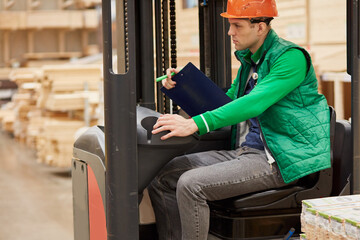 Fototapeta na wymiar side view on caucasian warehouse worker sitting inside of cab, wearing green unifrom and protective helmet, man make notes connected with work