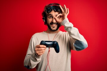 Young gamer man with curly hair and beard playing video game using joystick and headphones with happy face smiling doing ok sign with hand on eye looking through fingers