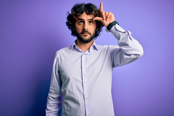 Young handsome business man with beard wearing shirt standing over purple background making fun of people with fingers on forehead doing loser gesture mocking and insulting.