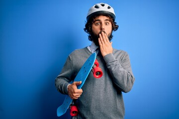 Handsome skater man with beard wearing security helmet holding skate over blue background cover mouth with hand shocked with shame for mistake, expression of fear, scared in silence, secret concept © Krakenimages.com