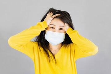 Young Asian woman wearing medical face mask and yellow shirt her hand on the head. isolated on gray...