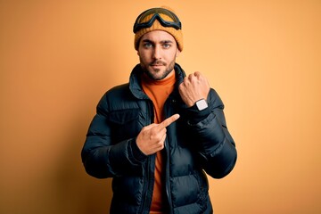 Young handsome skier man with beard wearing snow sportswear and ski goggles In hurry pointing to...