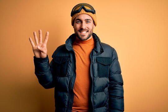 Young handsome skier man with beard wearing snow sportswear and ski goggles showing and pointing up with fingers number four while smiling confident and happy.