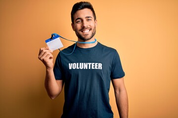 Handsome volunteer man with beard holding id card identification over yellow background with a happy face standing and smiling with a confident smile showing teeth