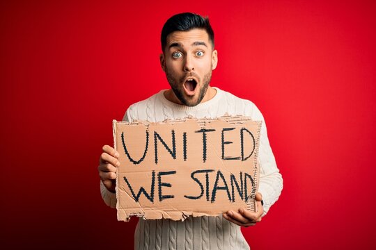 Young handsome activist man protesting holding cardboard with unity message scared in shock with a surprise face, afraid and excited with fear expression