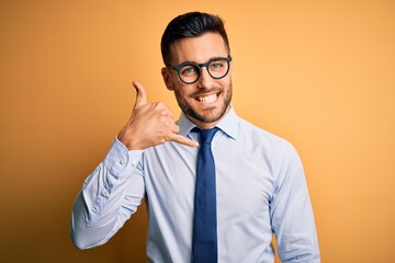 Young handsome businessman wearing tie and glasses standing over yellow background smiling doing phone gesture with hand and fingers like talking on the telephone. Communicating concepts.