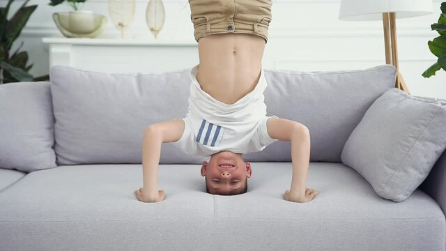 Front view of handsome sporty smiling teen boy which standing on his head and hands on the couch at home