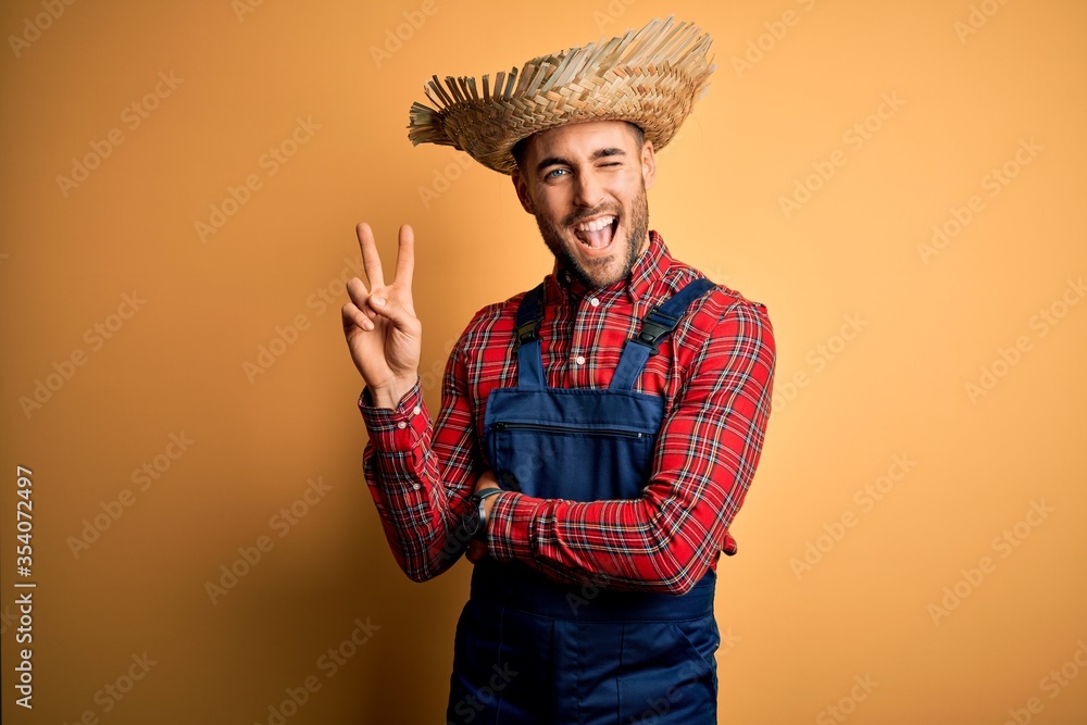 Wall mural Young rural farmer man wearing bib overall and countryside hat over yellow background smiling with happy face winking at the camera doing victory sign. Number two.