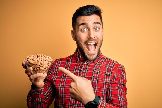 Young handsome man holding bowl with healthy peanuts over isolated yellow background very happy pointing with hand and finger
