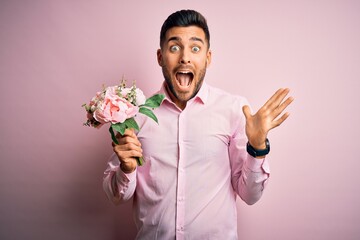 Young romatic man holding bouquet of spring flowers over pink isolatd background very happy and excited, winner expression celebrating victory screaming with big smile and raised hands