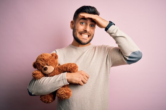 Young handsome man holding teddy bear standing over isolated pink background stressed with hand on head, shocked with shame and surprise face, angry and frustrated. Fear and upset for mistake.