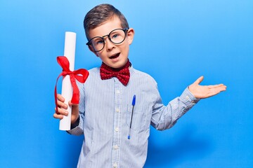 Cute blond kid wearing nerd bow tie and glasses holding diploma celebrating achievement with happy...