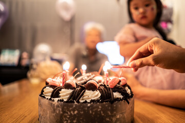 Group of diversity people in big family, young children and old parents having fun celebrate happy birthday anniversary party together at home. Woman is lighting candles on ice cream cake on table.