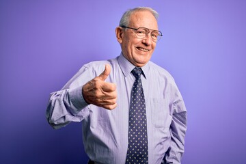 Grey haired senior business man wearing glasses standing over purple isolated background doing happy thumbs up gesture with hand. Approving expression looking at the camera showing success.