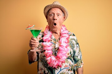 Grey haired senior man wearing summer hat and hawaiian lei drinking a cocktail scared in shock with...