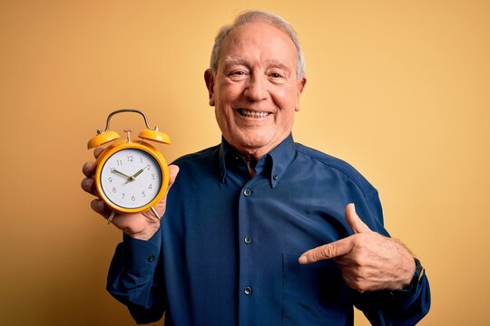 Senior grey haired man holding vintage alarm clock over yellow background with surprise face pointing finger to himself