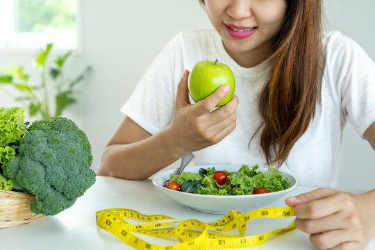 A Happy Woman Eating An Apple With Vegetable Salad For Health. Slim Woman Sitting On The Table With Fresh Raw Vegetables. Select Foods Have High Fiber Is Good For The Digestive System And Skin.