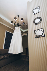 A beautiful white wedding dress of the bride hangs on a large chandelier in the interior. Photography, concept.