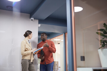 Wide angle view at two young business people chatting and gesturing while leaning against wall in contemporary office interior, copy space