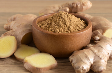 Ginger powder in a brown wooden bowl and fresh ginger root on a wooden background.