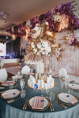 Banquet table with blue velvet tablecloth, candles, Alexa Dining Chair. High stand with a composition of rose flowers, hydrangeas, golden leaves. Against the background of golden racks with flowers, s