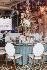 Banquet table with blue velvet tablecloth, candles, Alexa Dining Chair. High stand with a composition of rose flowers, hydrangeas, golden leaves. Against the background of golden racks with flowers, s