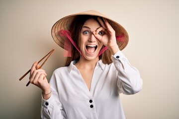 Young beautiful redhead woman wearing asian traditional hat holding wooden chopsticks with happy...