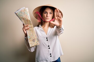 Young beautiful redhead woman wearing asian traditional hat holding city map with open hand doing stop sign with serious and confident expression, defense gesture