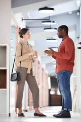 Full length side view portrait of two contemporary business people chatting while standing in office hall