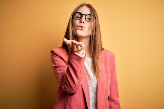 Young beautiful redhead woman wearing jacket and glasses over isolated yellow background looking at the camera blowing a kiss with hand on air being lovely and sexy. Love expression.