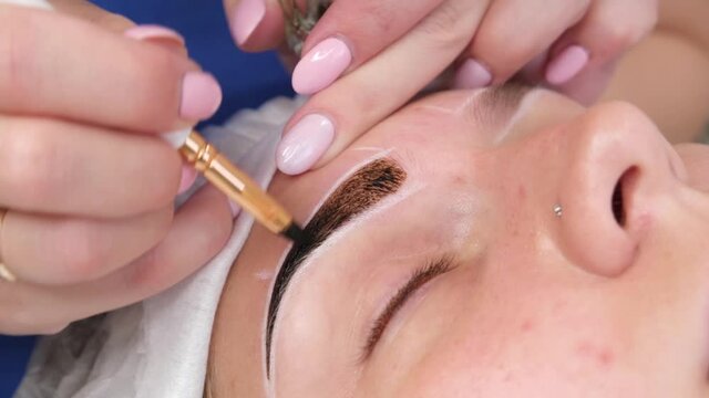 Permanent make-up for eyebrows of woman with thick brows in beauty salon. Closeup beautician doing coloring tattooing eyebrow.
