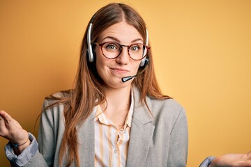 Young redhead call center agent woman overworked wearing glasses using headset clueless and confused expression with arms and hands raised. Doubt concept.