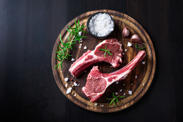 Two raw lamb chops on a wooden cutting board with spices on a dark wooden background