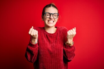 Young beautiful redhead woman wearing casual sweater over isolated red background very happy and excited doing winner gesture with arms raised, smiling and screaming for success. Celebration concept.