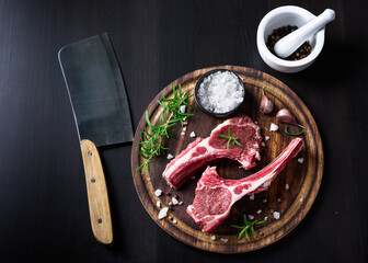 Two raw lamb chops on a wooden cutting board with spices on a dark wooden background