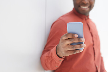 Close up of unrecognizable African-American man using smartphone while standing against while wall and smiling, focus on male hand holding phone in foreground, copy space