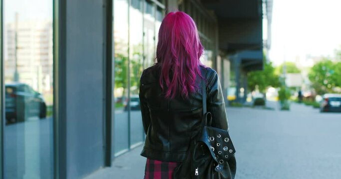 Rear of young Caucasian extraordinary girl with pink hair and in black leather jacket walking outdoor, then turning to camera. Back view on stylish female hipster strolling the street and turn back.