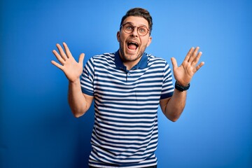 Young man with blue eyes wearing glasses and casual striped t-shirt over blue background celebrating crazy and amazed for success with arms raised and open eyes screaming excited. Winner concept