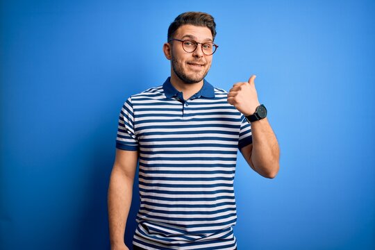 Young man with blue eyes wearing glasses and casual striped t-shirt over blue background smiling with happy face looking and pointing to the side with thumb up.