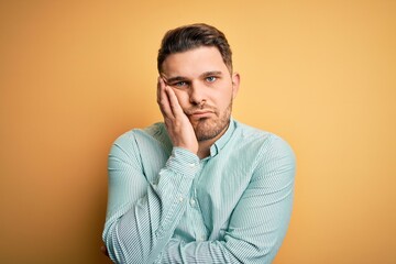 Young business man with blue eyes wearing elegant green shirt over yellow background thinking looking tired and bored with depression problems with crossed arms.