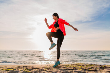 young smiling attractive slim woman doing sports in morning sunrise jumping on sea beach in sports...