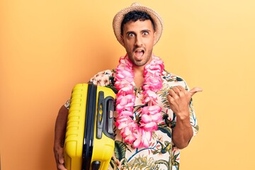 Young hispanic man wearing summer hat holding cabin bag pointing thumb up to the side smiling happy with open mouth