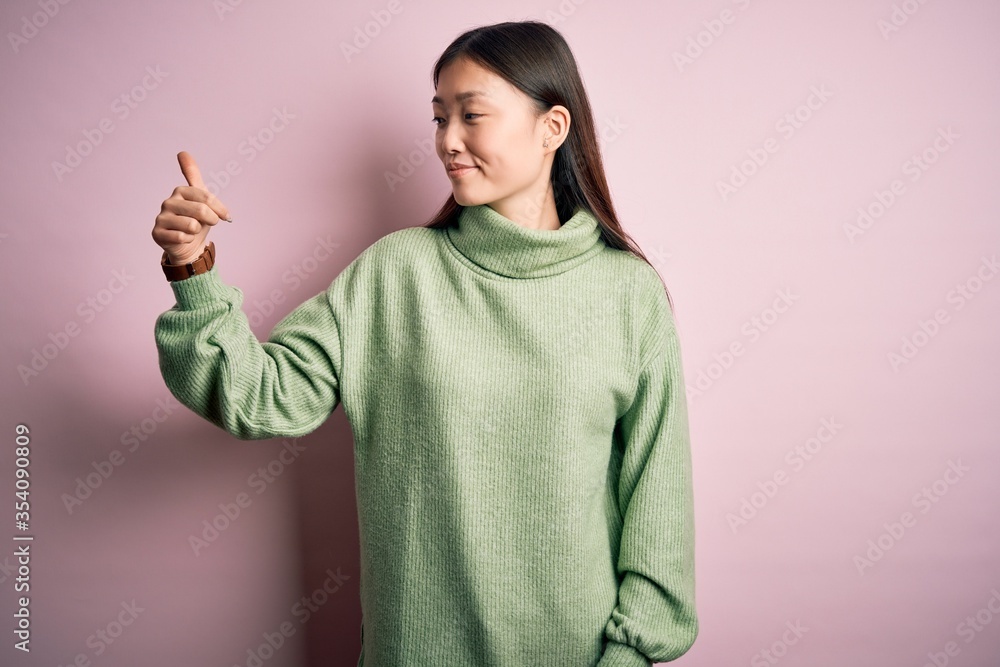 Poster Young beautiful asian woman wearing green winter sweater over pink solated background Looking proud, smiling doing thumbs up gesture to the side