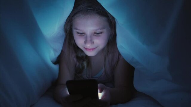 Alpha child leisure. Night social media. Girl reading fun content on smartphone under blanket before sleep.