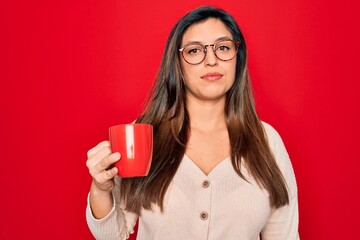 Young hispanic woman wearing glasses drinking a cup of coffee over red background with a confident expression on smart face thinking serious