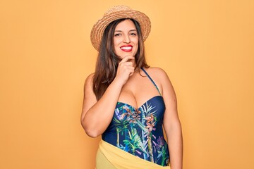 Young hispanic woman wearing summer hat and swimsuit over yellow background looking confident at the camera with smile with crossed arms and hand raised on chin. Thinking positive.