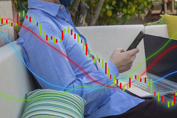 YOUNG MAN WITH BLUE SHIRT WORKING FROM HOME WITH HIS LAPTOP AND PHONE ON A SOFA.FINANCIAL CHART AND STOCK MARKET GRAPH