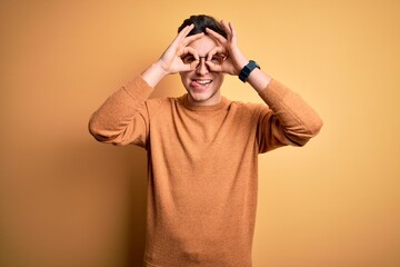 Young handsome caucasian man wearing glasses and casual winter sweater over yellow background doing ok gesture like binoculars sticking tongue out, eyes looking through fingers. Crazy expression.